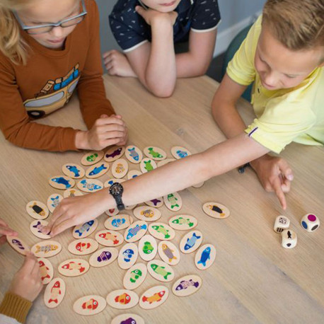 Nens juguen amb peces de fusta ovalades amb peixos acolorits i daus sobre una taula de to clar.