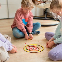 Nens asseguts a terra llancen petites fitxes de colors cap a un tauler circular de fusta amb cèrcols grocs i vermells.