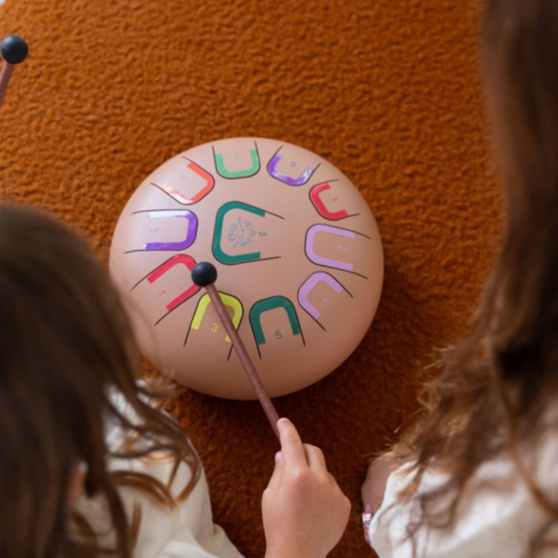 Tambor circular Tambú+ Deep Flow color nude de 25cm con 11 lengüetas de colores, tocado con una baqueta sobre fondo naranja.