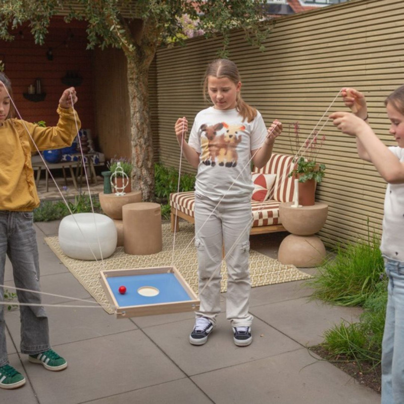 Juego cooperativo de madera con tablero azul y cuerdas, donde varios niños coordinan sus movimientos en un jardín exterior.