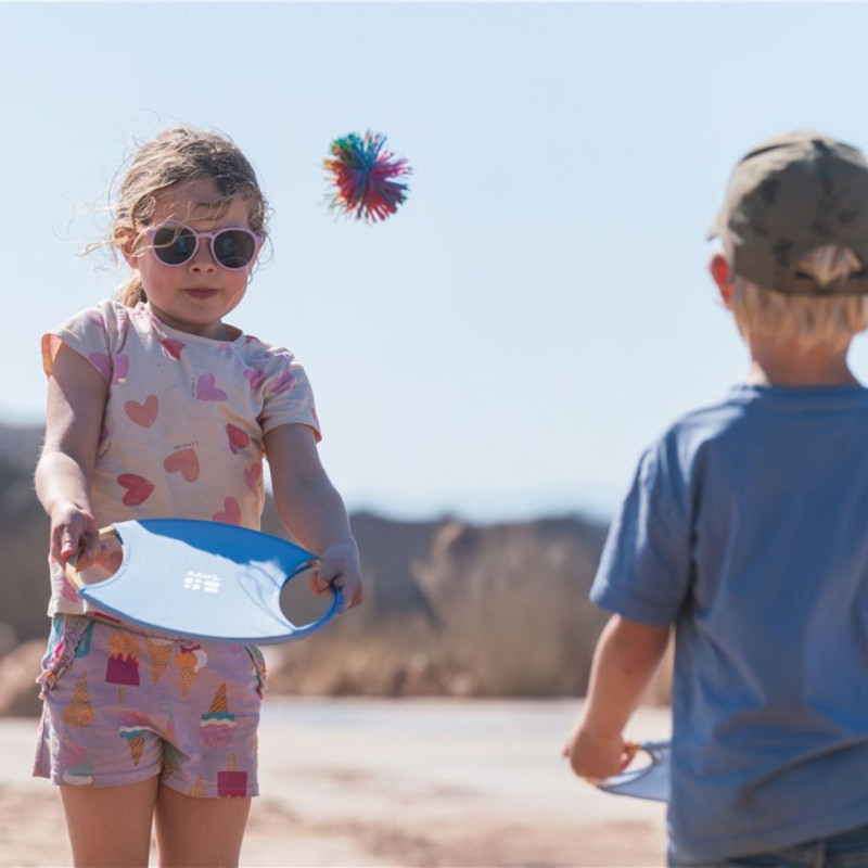 Dos niños juegan con raquetas circulares de bambú y borde azul lanzando una pelota de colores al aire.