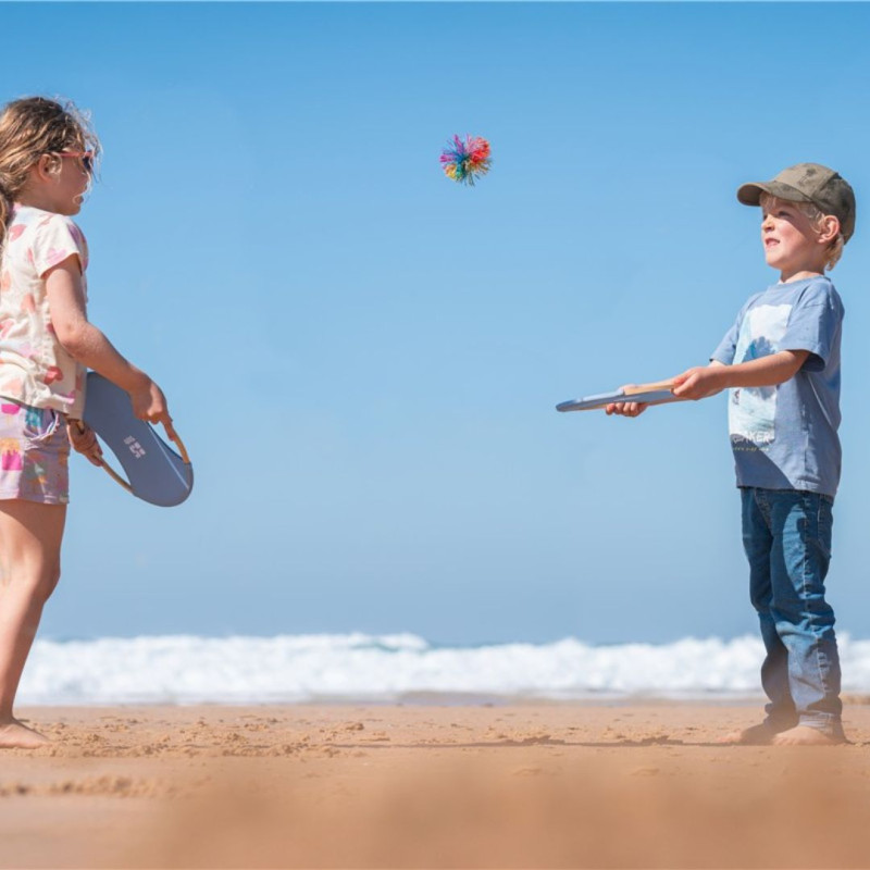 Niños jugando en la playa con un set de dos raquetas de bambú circulares y una pelota pequeña de colores vivos.