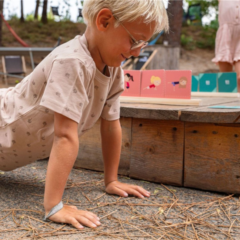 Niño imita una postura de ejercicio frente a tarjetas de colores rosa y azul del juego de memoria y poses al aire libre.