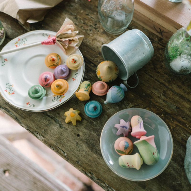 Flors de fusta artesanals en tons pastel, escampades sobre plats i una petita galleda metàl·lica sobre una taula rústica.