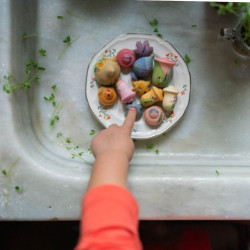 Flores de madera pintadas a mano en tonos pastel sobre un plato blanco, con la mano de un niño señalando las piezas.